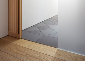 Wooden sliding door threshold detail. Chestnut wood floor with hand-carved surface treatment (Naguri), handmade dark gray floor tiling in background, manufactured in Nara, Japan.
